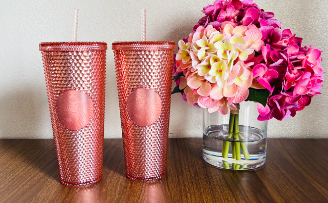 Two Rose Gold Starbucks Tumblers Next to a Vase with Flowers on a Table